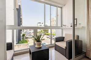 A balcony with a view of a parking lot and buildings, a potted plant, wicker furniture, and a glass door. - Modern 2BR Apartment Near Agora Mall (Santo Domingo)