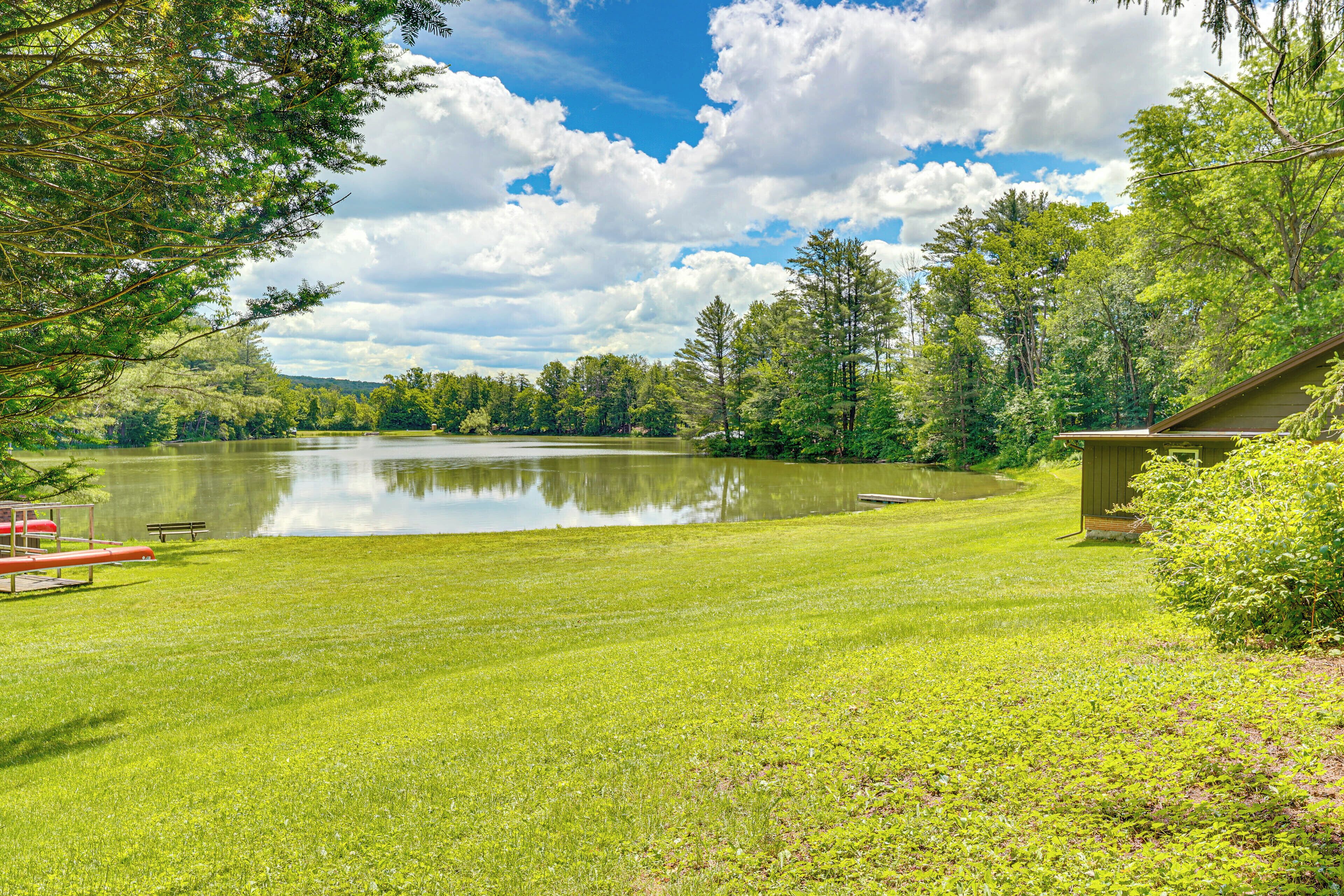 A tranquil lakeside setting with a grassy lawn, a wooden dock, and a small building, likely a cabin or guesthouse.
