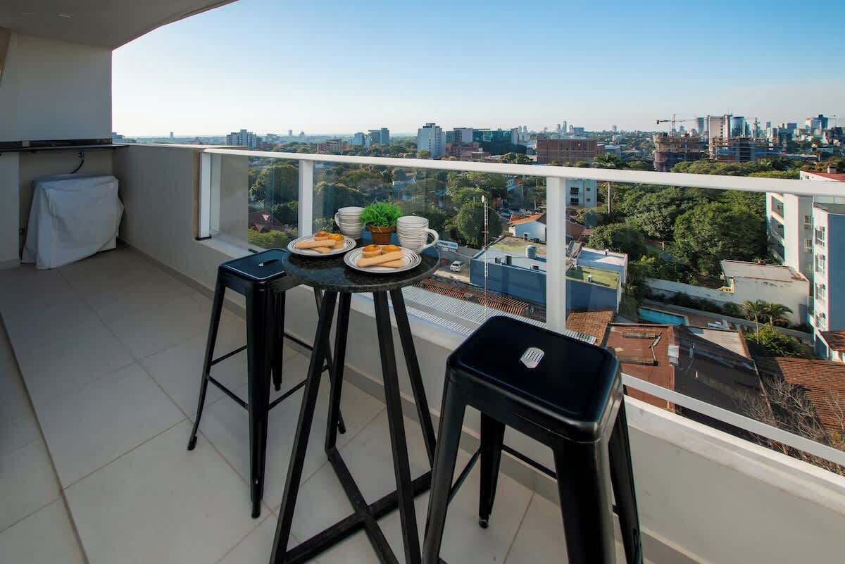 Un balcón con una mesa dispuesta para dos personas, una silla y una planta pequeña.