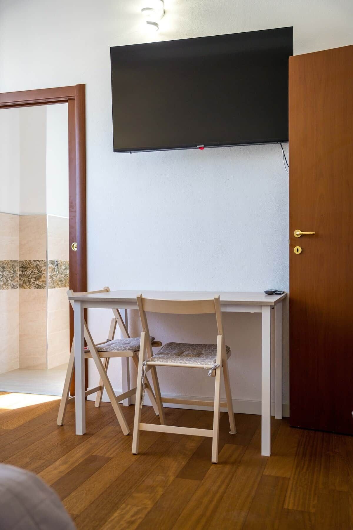 Una habitación con un escritorio de madera, dos sillas, un televisor montado en la pared y una puerta de madera.