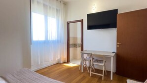 Quarto de hotel com cama, escrivaninha, cadeira, televisão e porta.