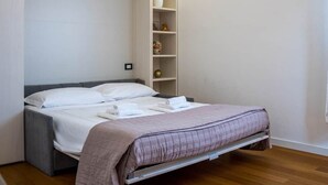 Quarto com cama, mesa de cabeceira, cadeira, prateleira com objetos decorativos e um aparelho de ar condicionado.