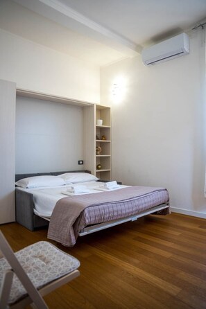 A bedroom with a bed, a bedside table, a chair, a shelf with decorative items, and an air conditioning unit. - Casa Helios Cozy Penthouse (Sassari)