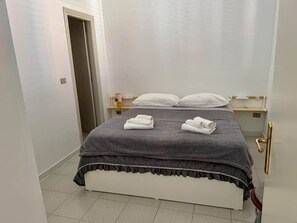 Ein ordentlich bezogenes Bett mit grauer Decke und zwei aufgerollten Handtüchern, ein Nachttisch mit Lampe und eine Tür mit goldener Klinke.