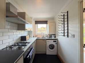 A compact kitchen with a gas stove, oven, washing machine, and a window with a view of buildings. - Lovely 2BDR Flat with Balcony in Vibrant Islington (Greater London)