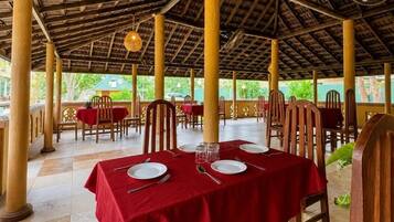 Un área de comedor al aire libre con mesas y sillas de madera, techo de paja y vistas a la vegetación.