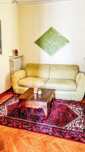 Una sala de estar con sofá, mesa de centro, radiador, alfombra decorativa y una obra de arte en forma de rombo colgada en la pared.