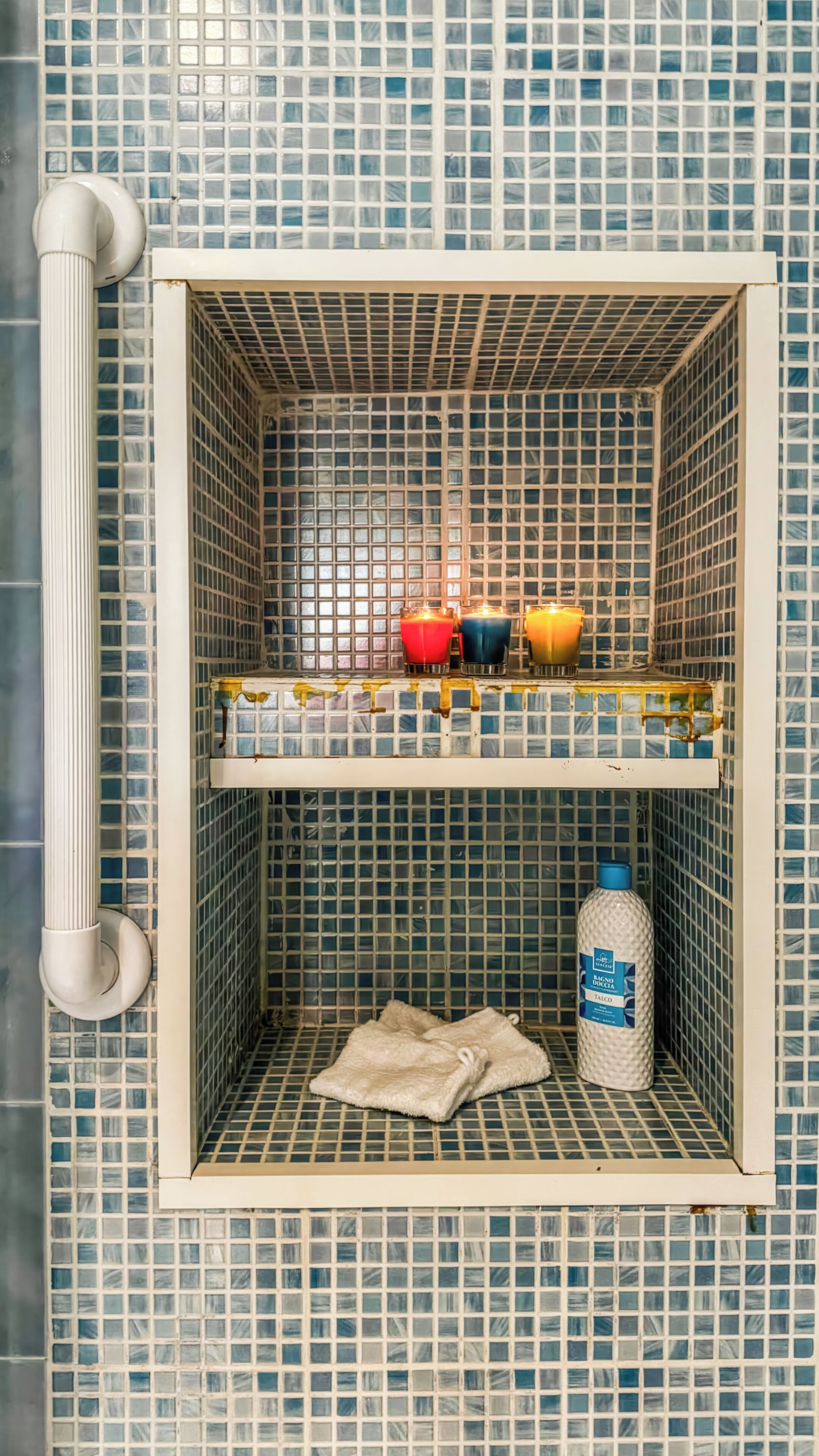 A bathroom with a tiled wall, a shelf with candles, a bottle of hand soap, and a towel.