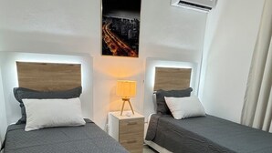 Ett hotellrum med två sängar, en takfläkt, en luftkonditioneringsanläggning, ett nattduksbord med en lampa och en tavla med en stadsbild på väggen.