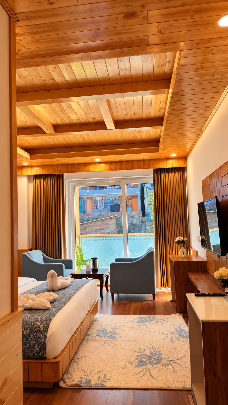 A hotel room with a wooden ceiling, a bed with a patterned bedspread, two armchairs, a small table with a vase of flowers, and a television.