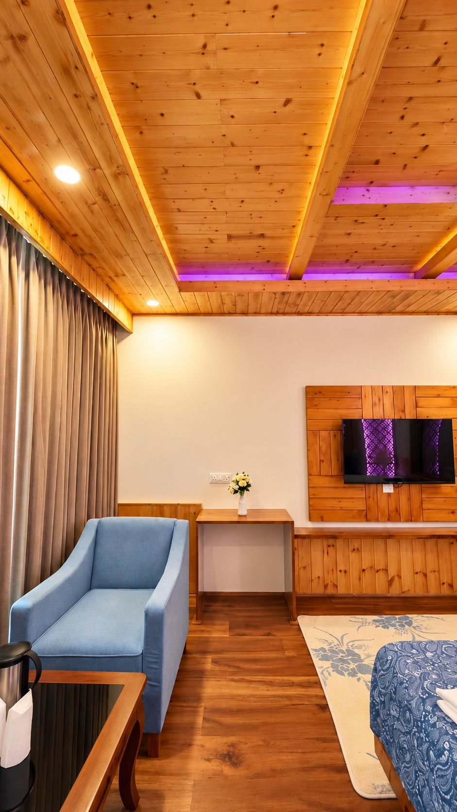 A room with a wooden ceiling, a blue sofa, a wooden table with a kettle, a TV on a wooden wall, and a patterned rug.