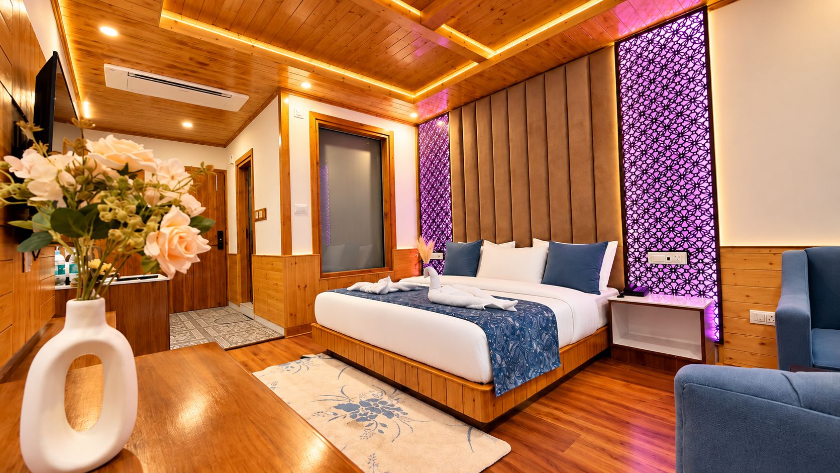 A modern hotel room with a wooden floor, a bed with a blue bedspread, a bedside table, a chair, and a decorative wall panel.