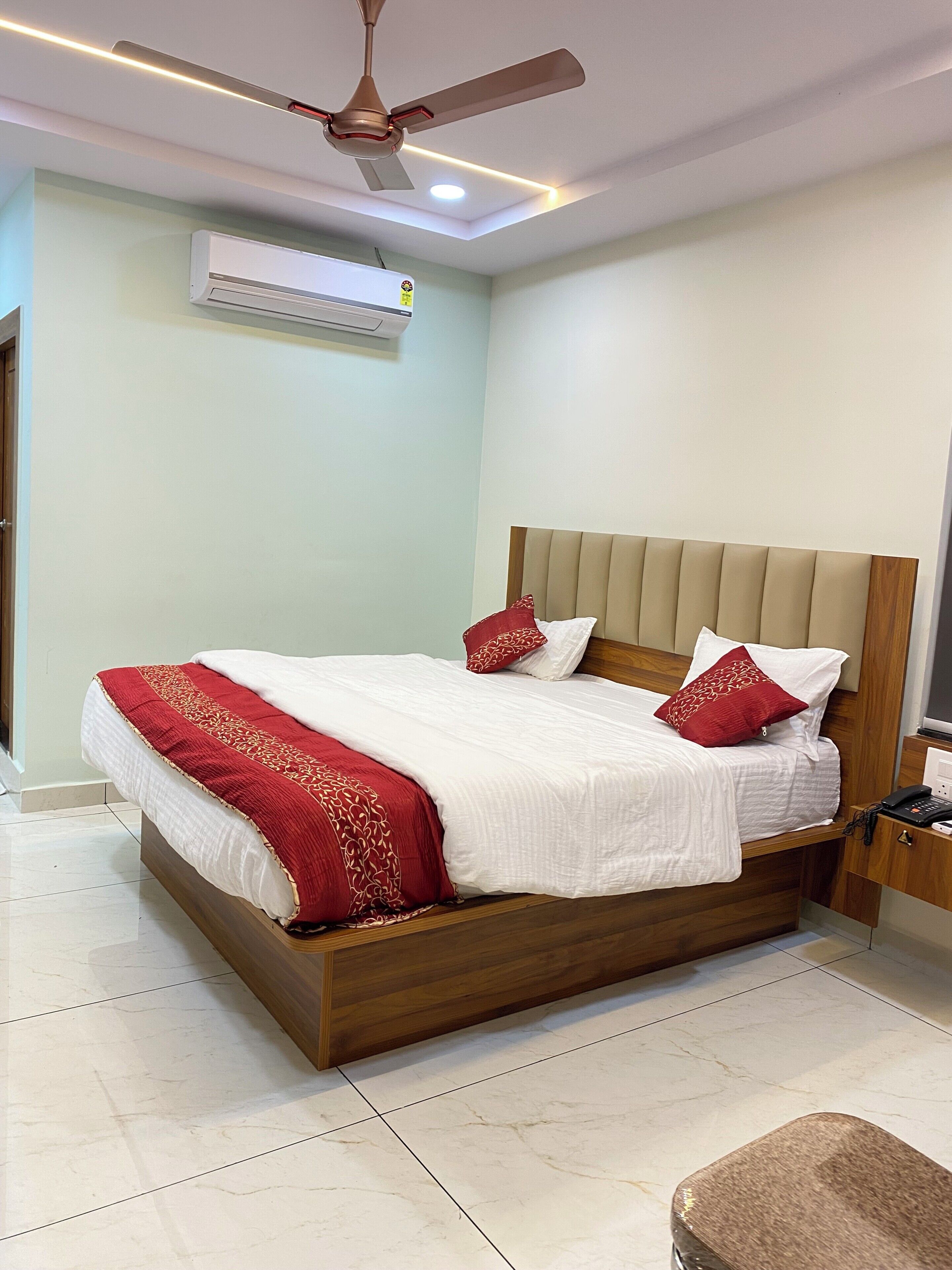 A hotel room with a bed, a ceiling fan, an air conditioning unit, a bedside table with a phone, and a nightstand.