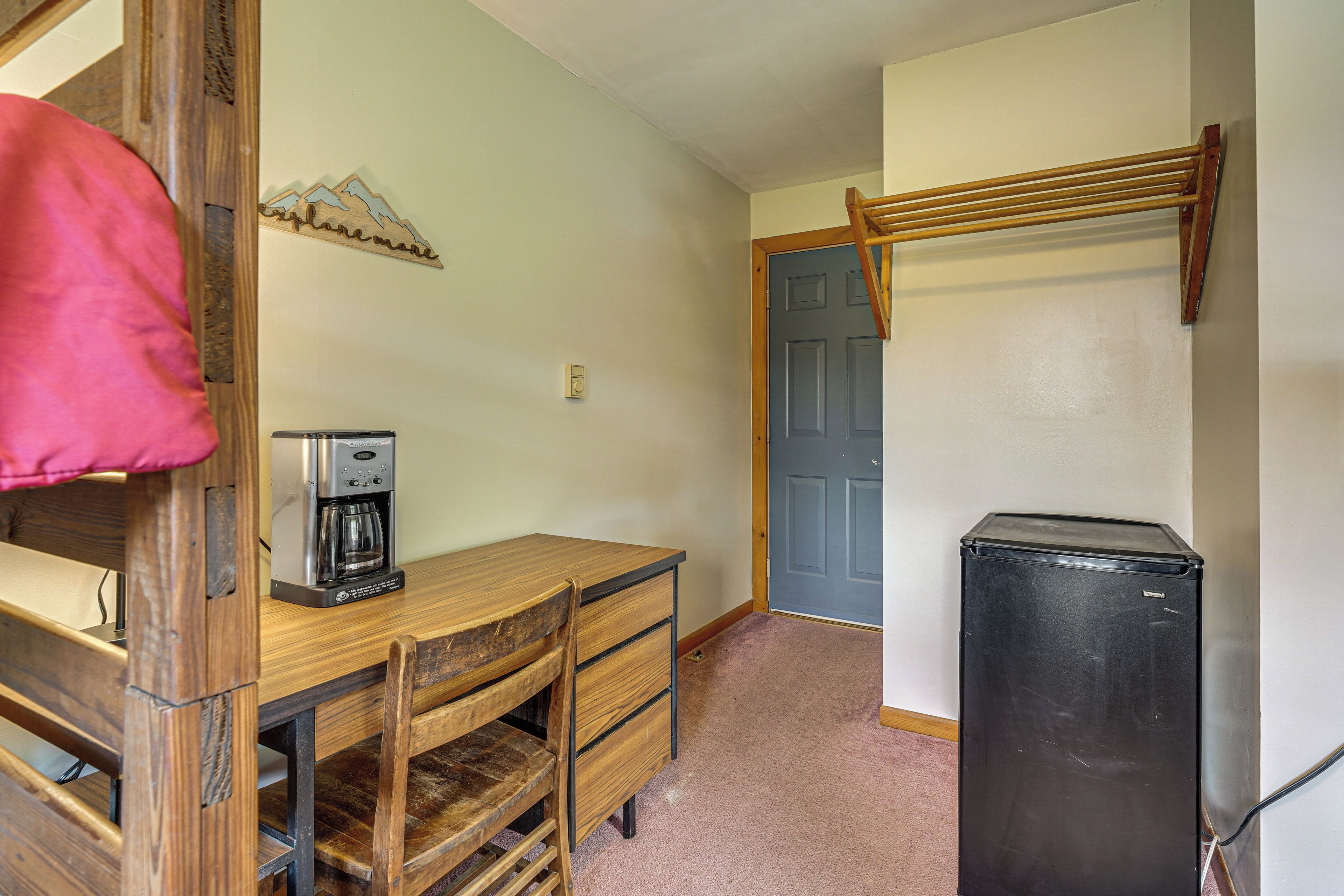A compact room with a wooden desk, a coffee maker, a refrigerator, and a door with a wooden ladder.