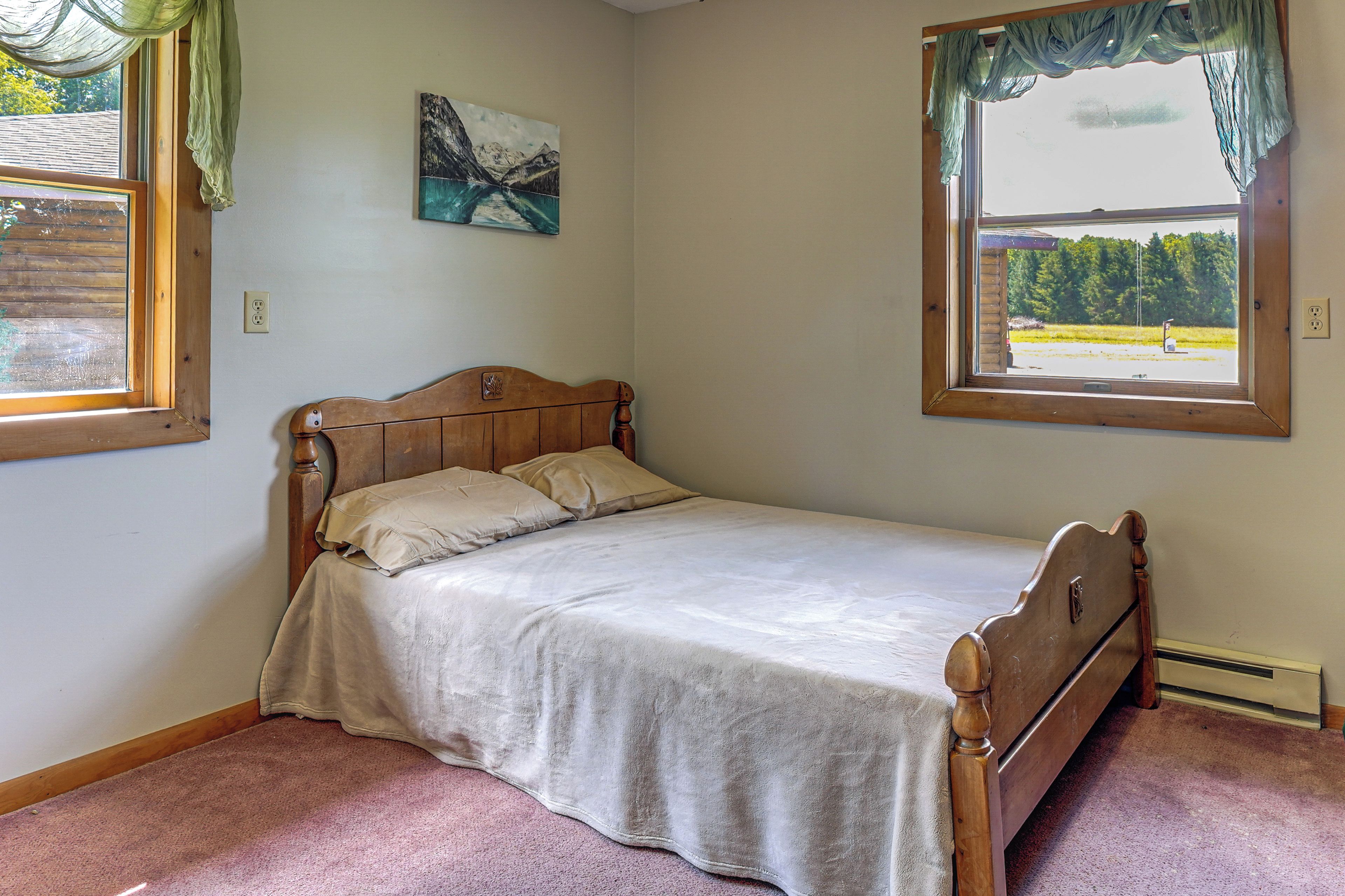 A single bed with a wooden headboard and a plain bedspread, a painting on the wall, and a window with a view of a landscape.