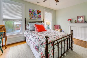 Ein Schlafzimmer mit einem Bett, einem Holzkopfstück, einem Fenster mit Jalousien, einem Bild an der Wand und einem Kaminvorsatz mit Spiegel und Blumen.