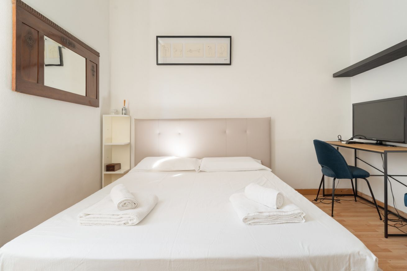 A hotel room with a bed, a desk, a chair, a TV, a mirror, and a framed picture on the wall.