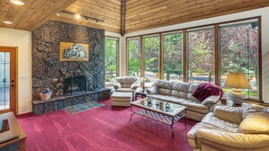A spacious living room with a stone fireplace, leather furniture, and large windows offering a view of trees. - Fire Pit & Wildlife Views! Tranquil Bend Retreat (Bend)