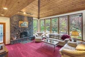 A spacious living room with a stone fireplace, leather furniture, and large windows offering a view of trees. - Fire Pit & Wildlife Views! Tranquil Bend Retreat (Bend)