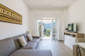 A modern living room with a grey sofa, a flat-screen TV, and a sliding glass door leading to a balcony with outdoor furniture. - GuestHost - Cozy Apartment with Balcony (Como)