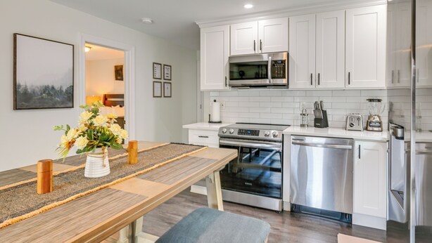 A modern kitchen with white cabinets, stainless steel appliances, and a wooden dining table with a vase of flowers. - Walk to Train! Updated Apt in Huntington Station (Huntington Station)