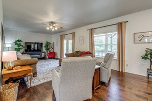 A spacious living room with a flat-screen TV, comfortable seating, a ceiling fan, and large windows with curtains. - 8 Mi to Bentonville: Serene Retreat on 8 Acres (Pea Ridge)