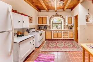 A kitchen with white appliances, wooden cabinets, a patterned rug, and a window with a view. - Traditional Taos Adobe Casita: Comfortable & Quiet (Taos)