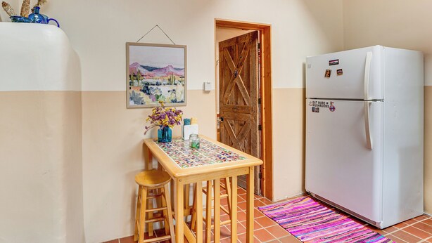 A kitchen with a wooden table, stools, a refrigerator, a door, and a tiled floor. - Traditional Taos Adobe Casita: Comfortable & Quiet (Taos)