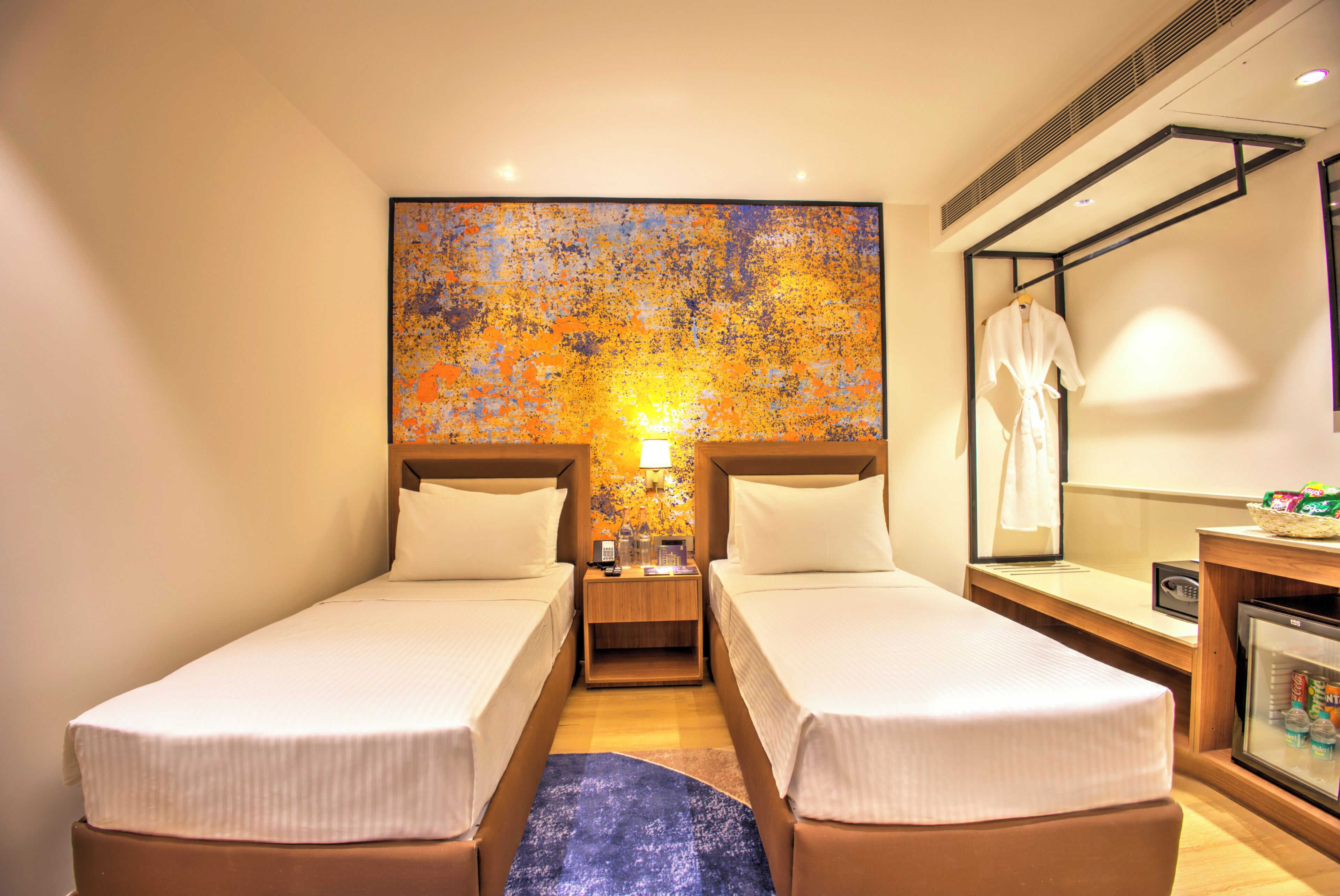 Quarto de hotel com duas camas, uma mesinha, uma mesinha de cabeceira, um espelho e um frigorífico.