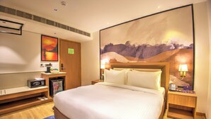 A hotel room with a large bed, a nightstand with a lamp, a small desk with a computer, a microwave, and a refrigerator. - Cozzet Yashobhoomi Dwarka (New Delhi)