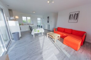 A modern living room with a red sofa, a wooden coffee table, and a dining area with a white table and chairs. - LA MAISON DES DUNES, sleeps 7, near the sea (Les Moitiers-d'Allonne)