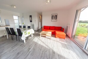 Un salon moderne comprenant un coin repas, un canapé avec des coussins orange et une grande fenêtre donnant sur l’extérieur.