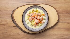 Een kom soep met zalm en groenten op een houten plank.