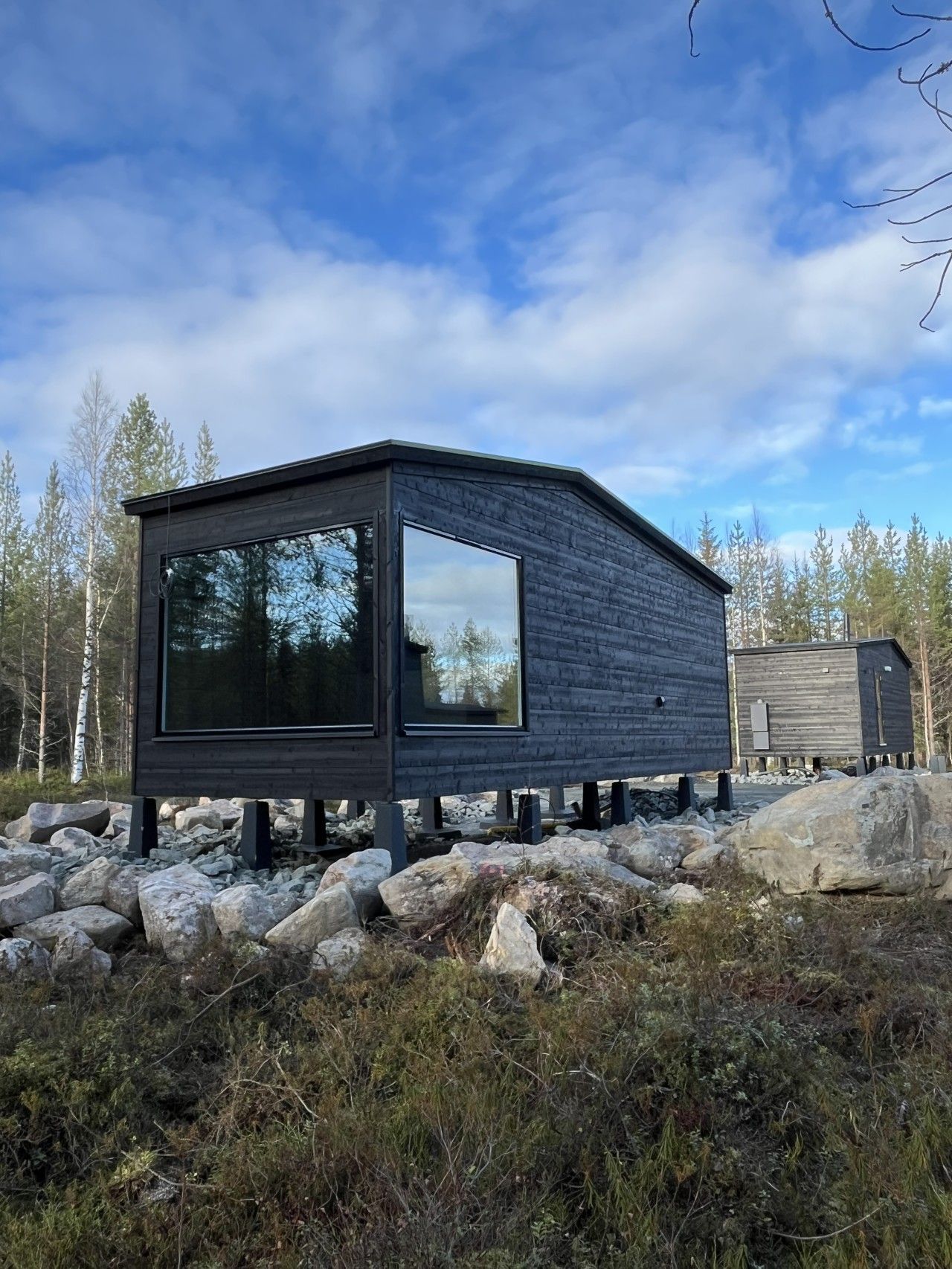 Eine moderne Holzhütte mit großem Fenster, gelegen auf felsigem Gelände mit Bäumen im Hintergrund.