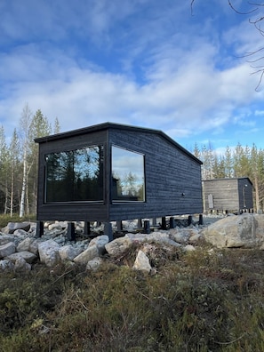 Une cabane moderne en bois, dotée d’une grande fenêtre, située sur un terrain rocheux, avec des arbres en arrière-plan.