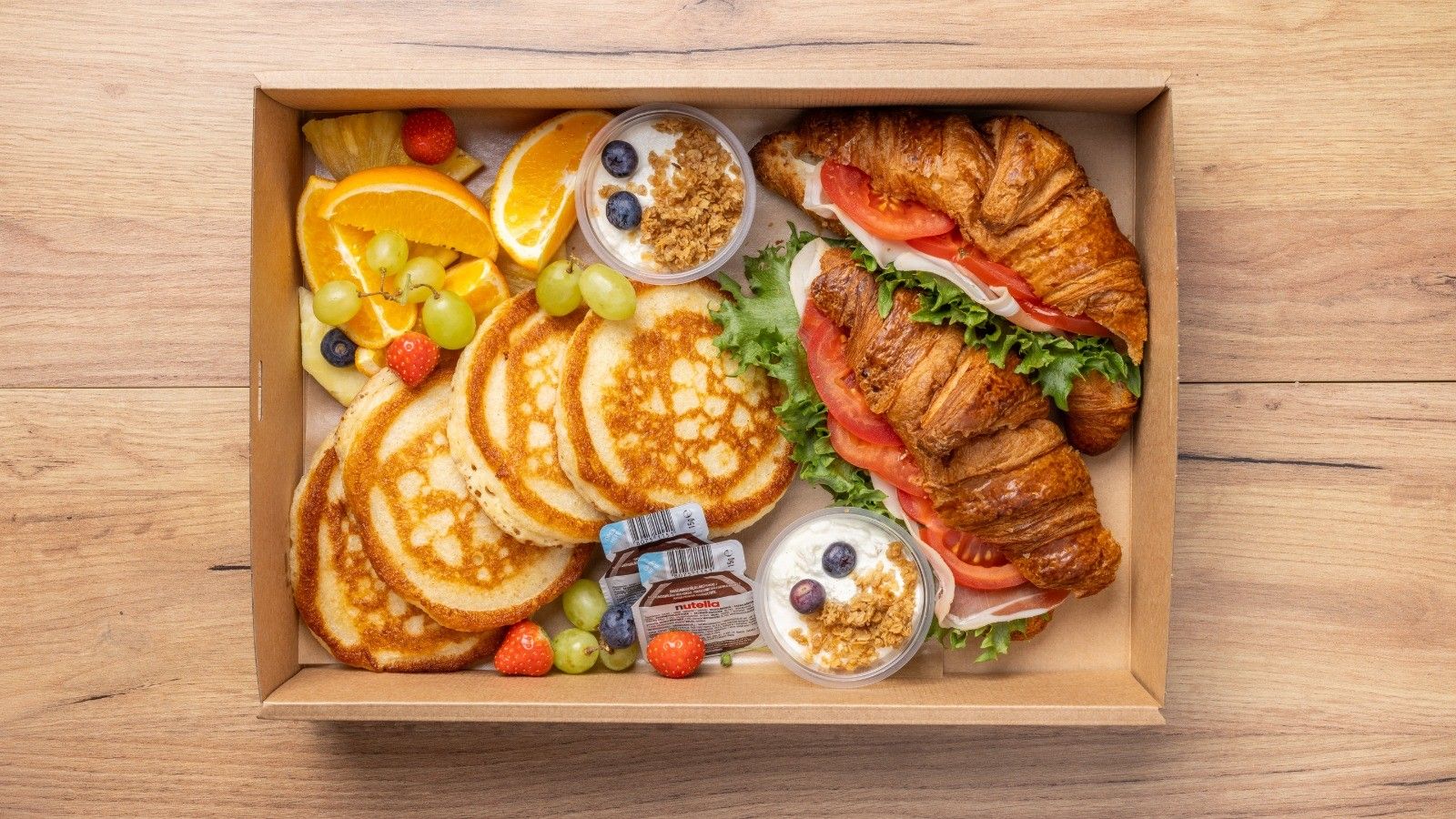 A breakfast box containing pancakes, croissants, fresh fruit, and yogurt.
