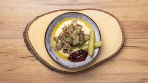 Ein Teller Essen mit Fleisch, Essiggurken und einer cremigen Soße auf einem Holztisch.