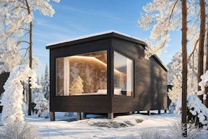 Une cabane moderne en bois, dotée d’une grande fenêtre en verre et entourée d’arbres couverts de neige.