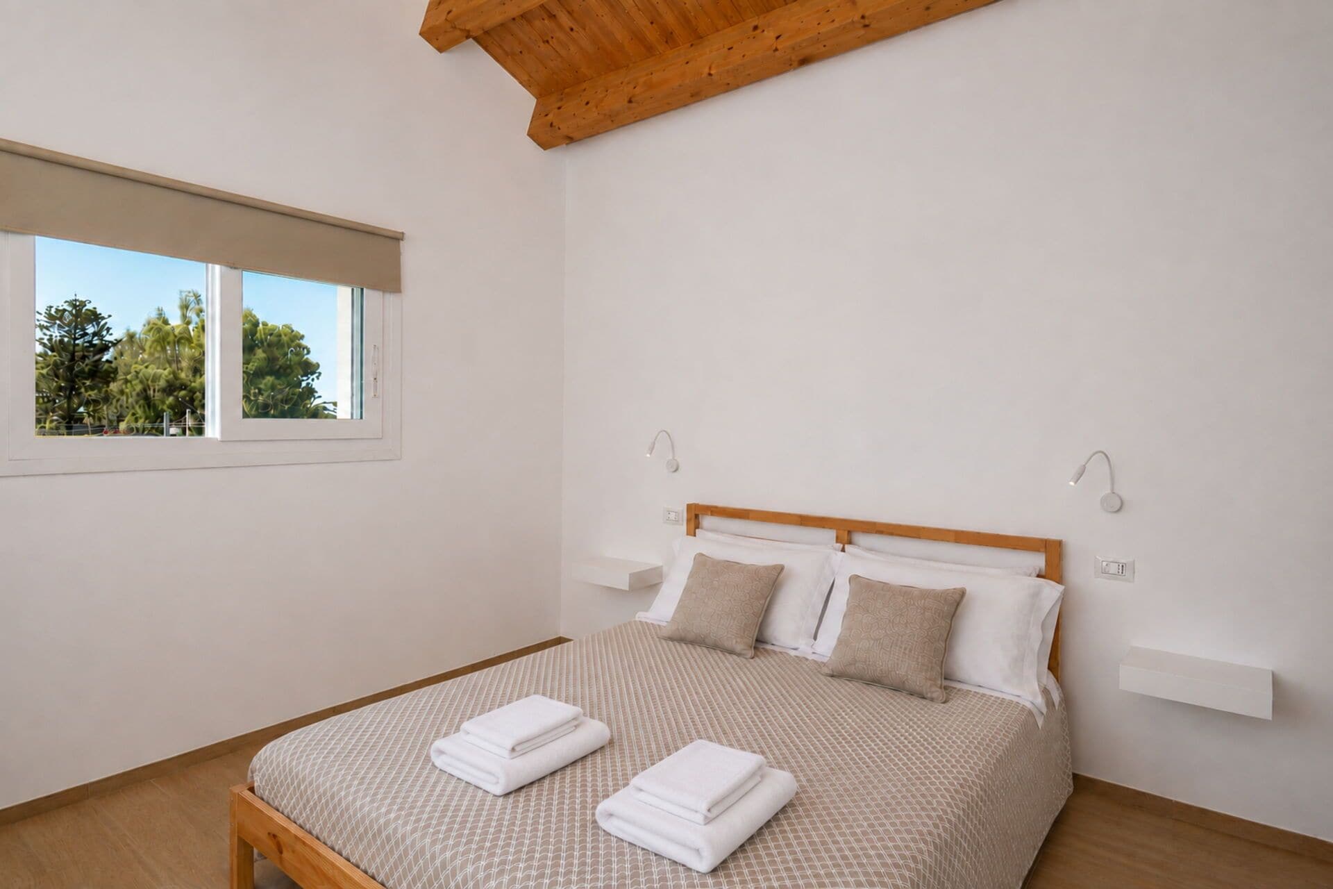 A bedroom with a bed, two bedside tables, a window, and a ceiling with exposed beams.