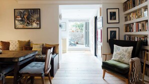 A room with a dining table, chairs, a sofa, a bookshelf, and a painting on the wall. - Farmhouse Chic Studio Flat with Private Garden (Greater London)