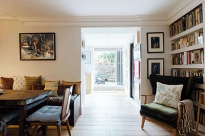 A room with a dining table, chairs, a sofa, a bookshelf, and a painting on the wall. - Farmhouse Chic Studio Flat with Private Garden (Greater London)