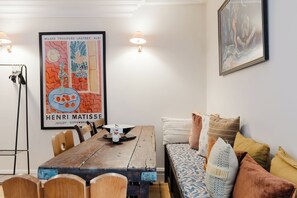 A dining area with a wooden table, chairs, and a bench with cushions. There are wall-mounted lights and a framed poster on the wall. - Farmhouse Chic Studio Flat with Private Garden (Greater London)