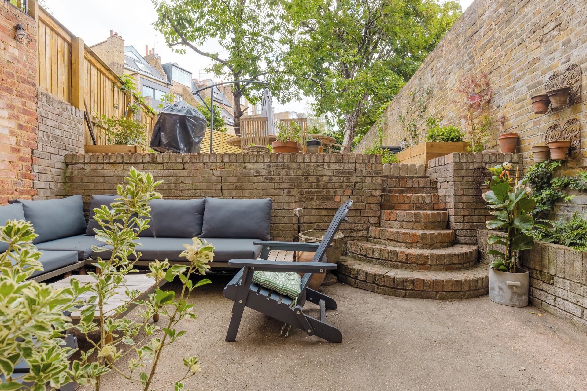 A courtyard with a brick wall, a wooden fence, a grey sofa, a lounge chair, and potted plants.