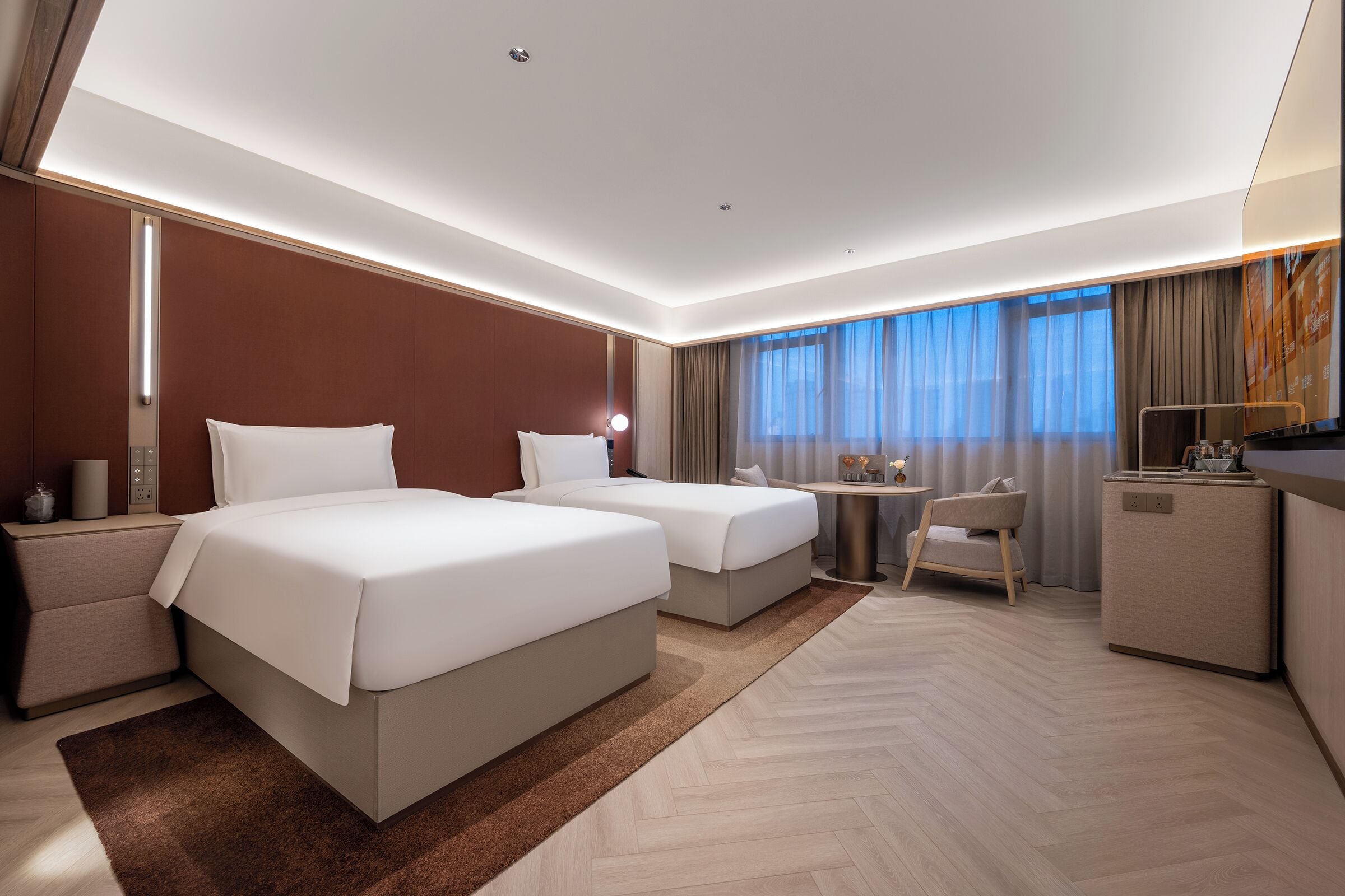 A hotel room with two beds, a desk, a chair, a minibar, and a large window with curtains.