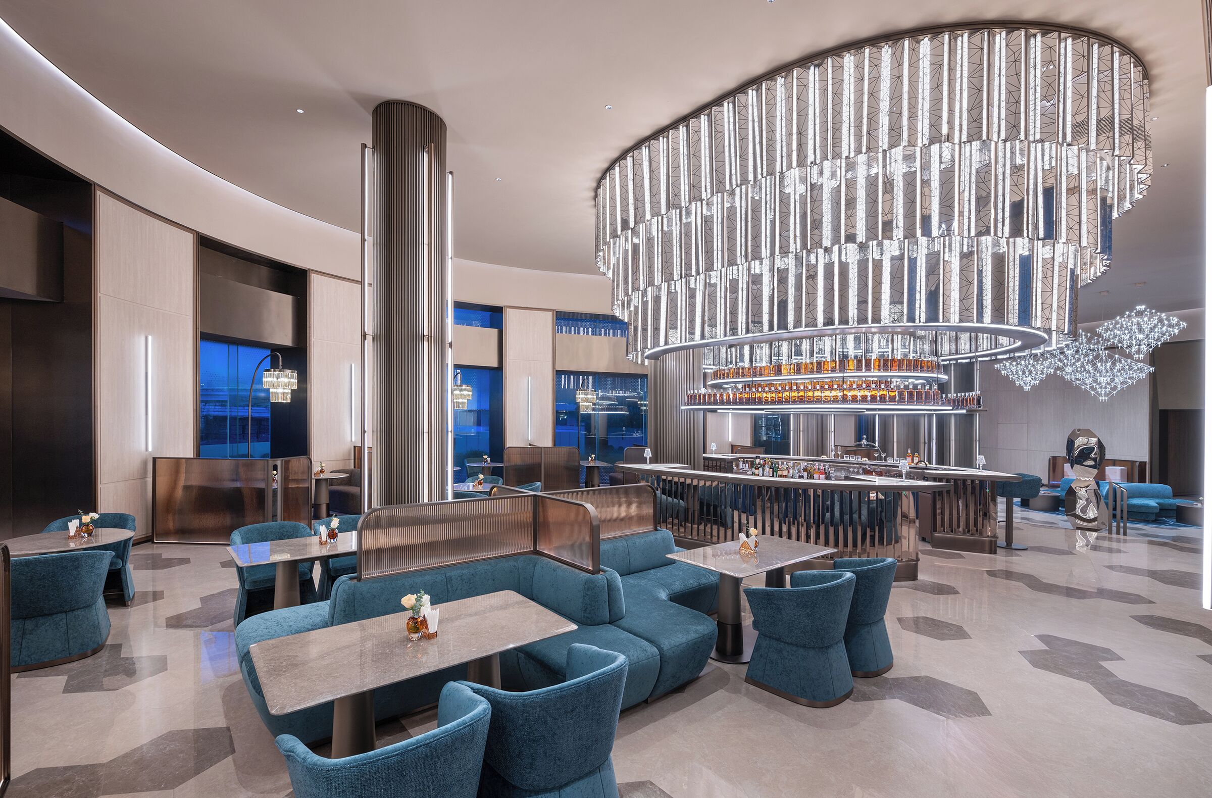 A modern hotel lobby with a central bar, seating areas, and a distinctive chandelier.