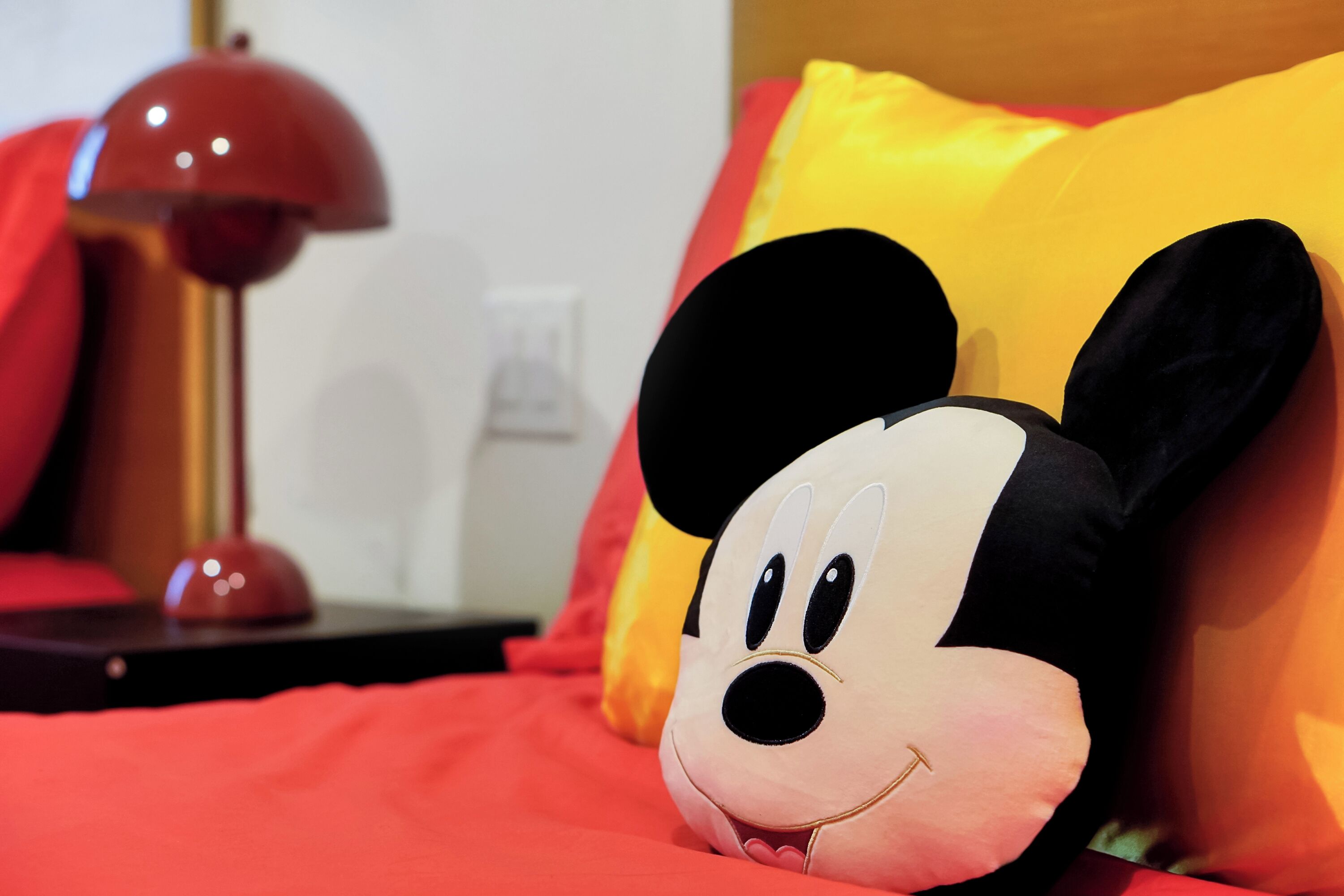 Een zacht Mickey Mouse kussen op een bed met een rode deken, een rode lamp en een geel kussen.