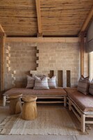 Una habitación rústica con techo de madera, pared de piedra y un banco con cojines. El suelo tiene dos taburetes de madera y una alfombra tejida.