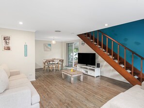 A living room with a wooden staircase, a dining area with a table and chairs, a television, and a sofa. - Boronia House in Sawtell Creekside Townhouse (Sawtell)