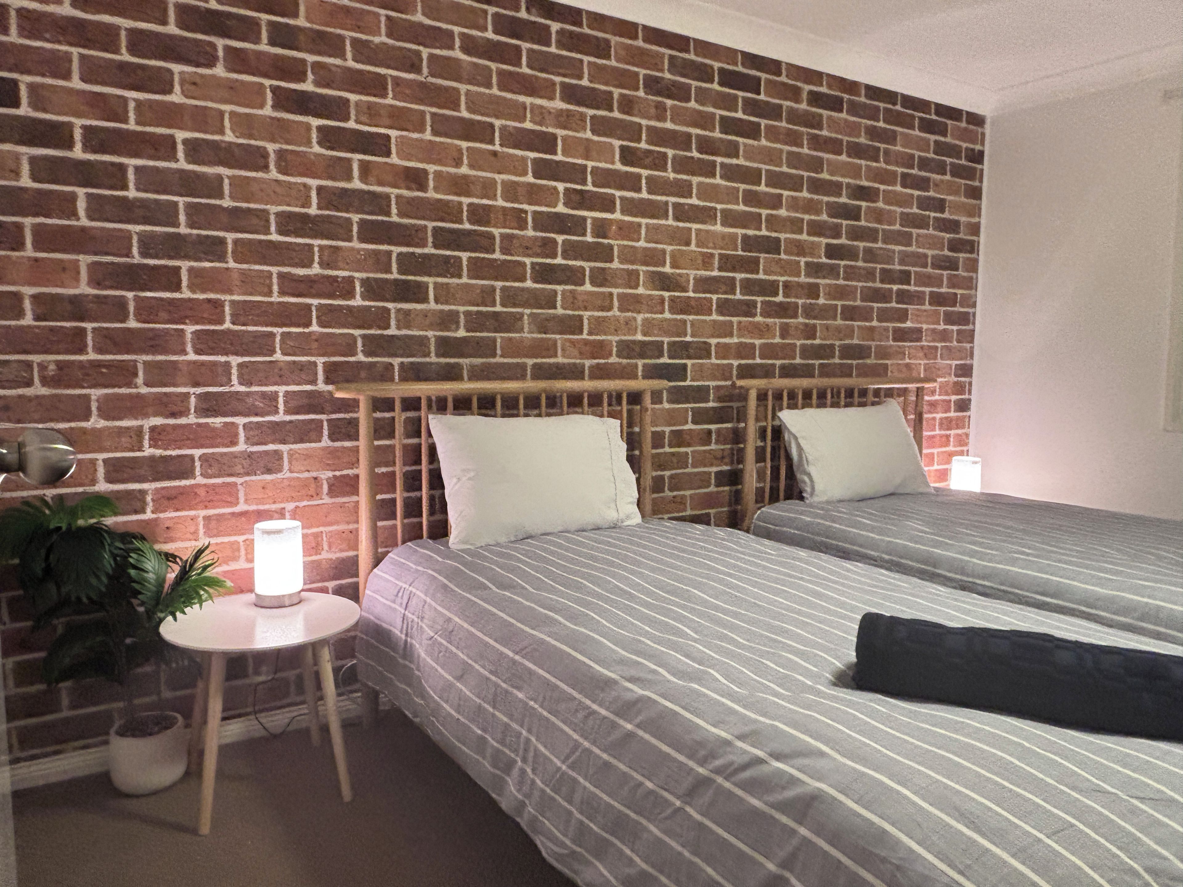 A room with two beds, a brick wall, a small table with a lamp, and a potted plant.