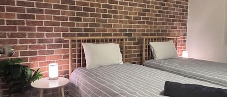 A room with two beds, a brick wall, a small table with a lamp, and a potted plant.